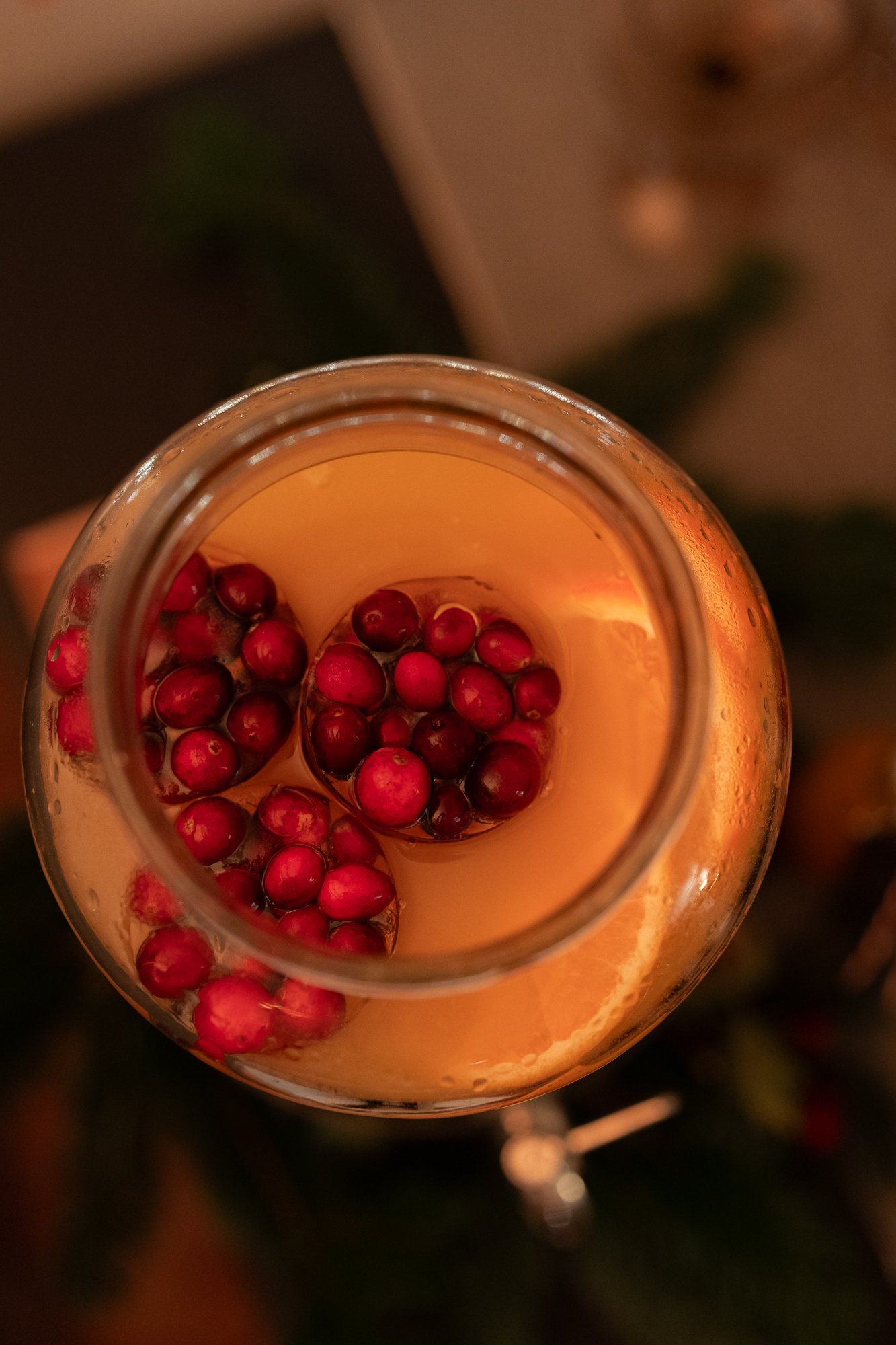 Vue de dessus d'un bocal en verre rempli d'une boisson claire, garni de tranches d'orange et de canneberges flottantes, avec des lumières tamisées en arrière-plan.