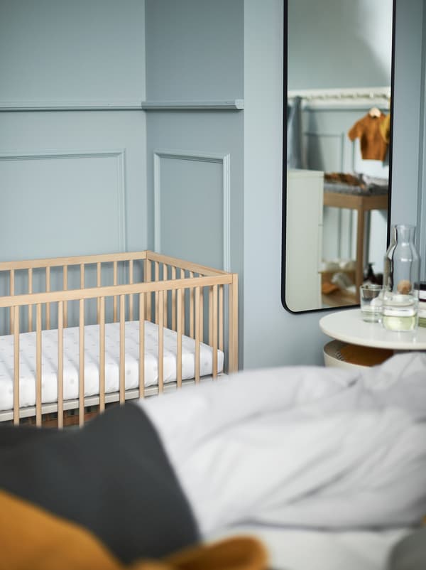 Vue partielle d’une chambre bleu clair avec une table de chevet blanche, un miroir et un lit bébé en bois SNIGLAR.
