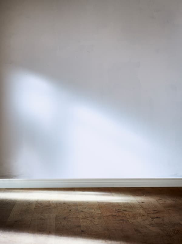 Vue partielle d’une pièce vide avec un mur blanc non peint et un sol en bois naturel. La lumière du soleil provient d’une fenêtre latérale.