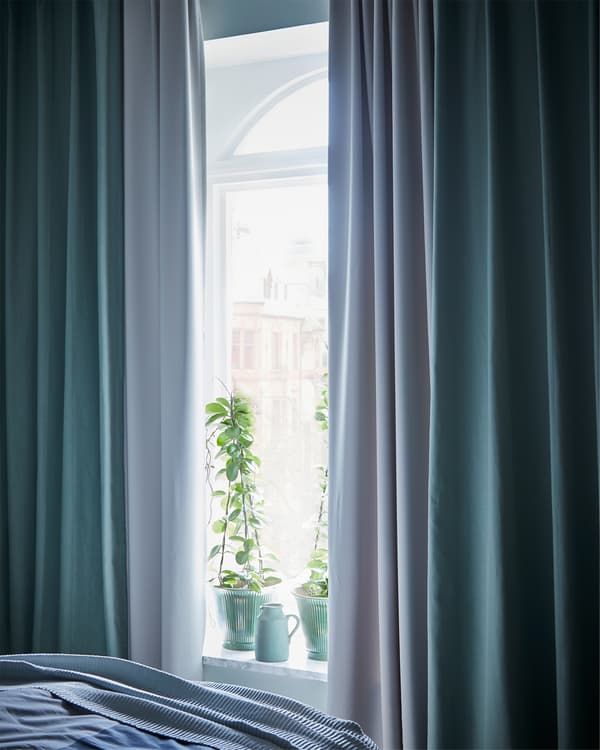 Vue sur l'extérieur par une fenêtre de chambre à coucher encadrée de rideaux gris clair et verts.