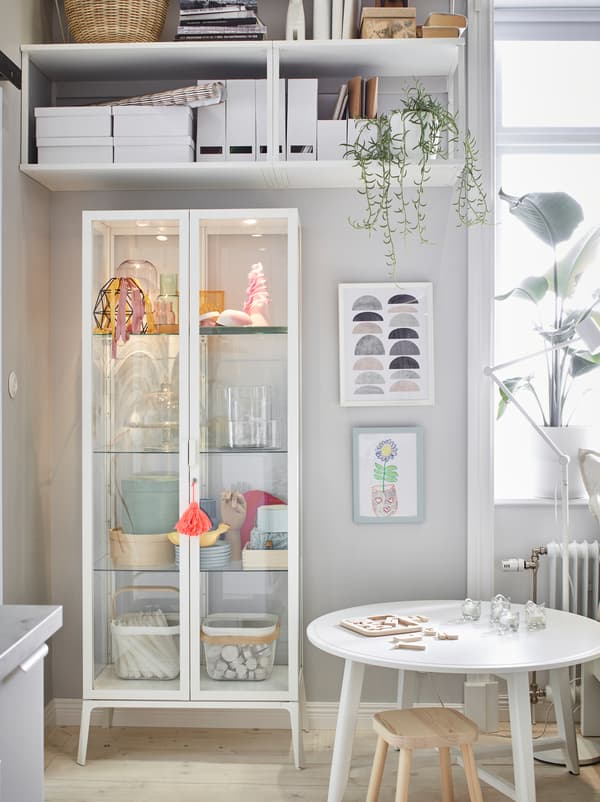 Vysoká biela vitrína MILSBO umiestnená v rohu pod nástennými policami s ďalším úložným priestorom.