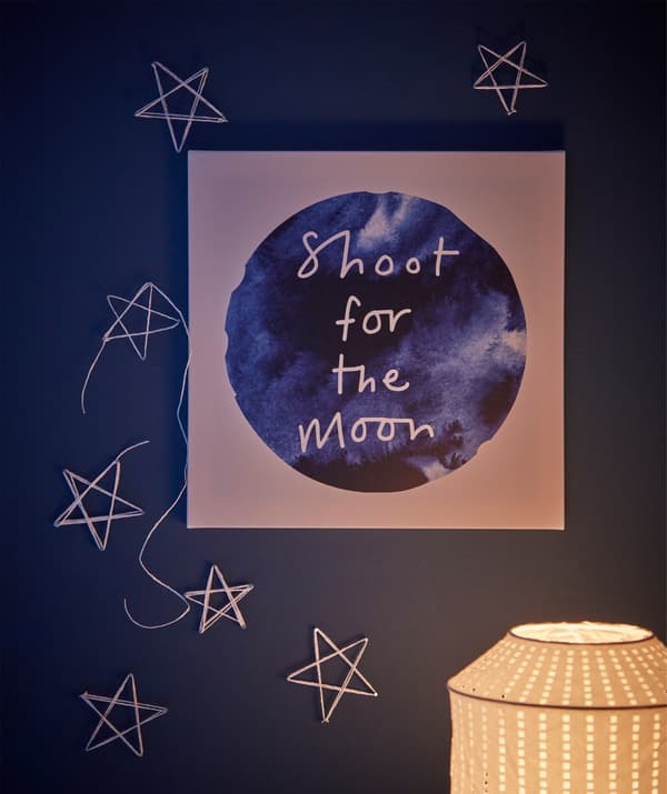 Wall above desk, adorned with stars made of ribbon and moon-themed PJÄTTERYD picture art reading 'Shoot for the moon'.