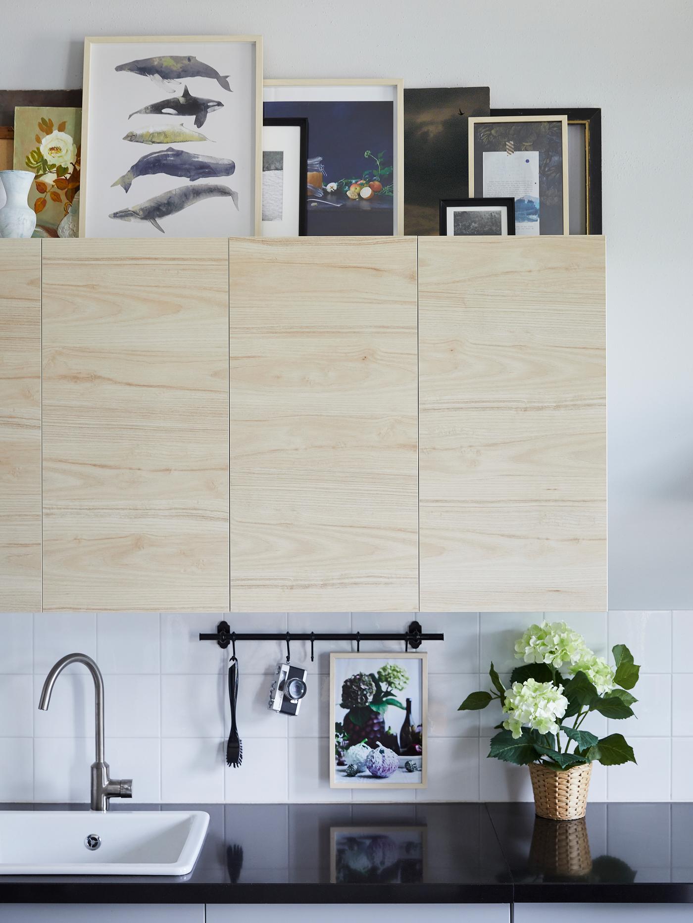 Wall cabinets with light ash wood-effect fronts. Framed prints are on top, a black worktop and a white sink are below.