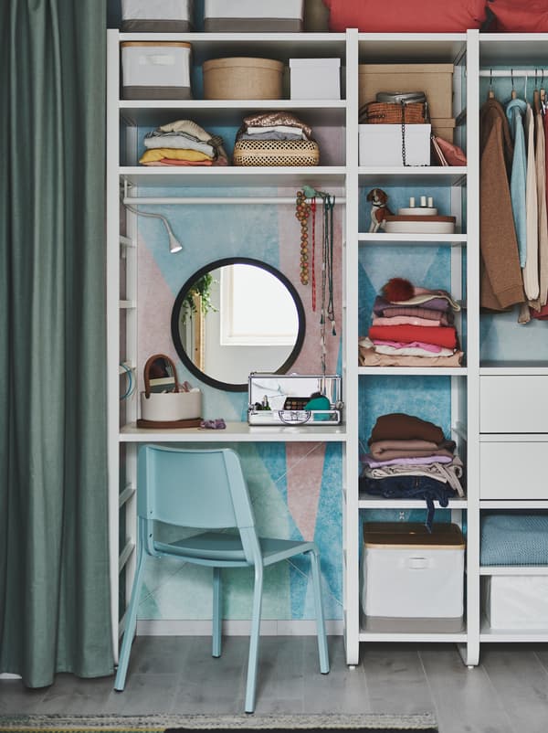 Wall-covering shelving unit used as a wardrobe, part of it acting as dressing table, with mirror, a TEODORES chair, and make-up.