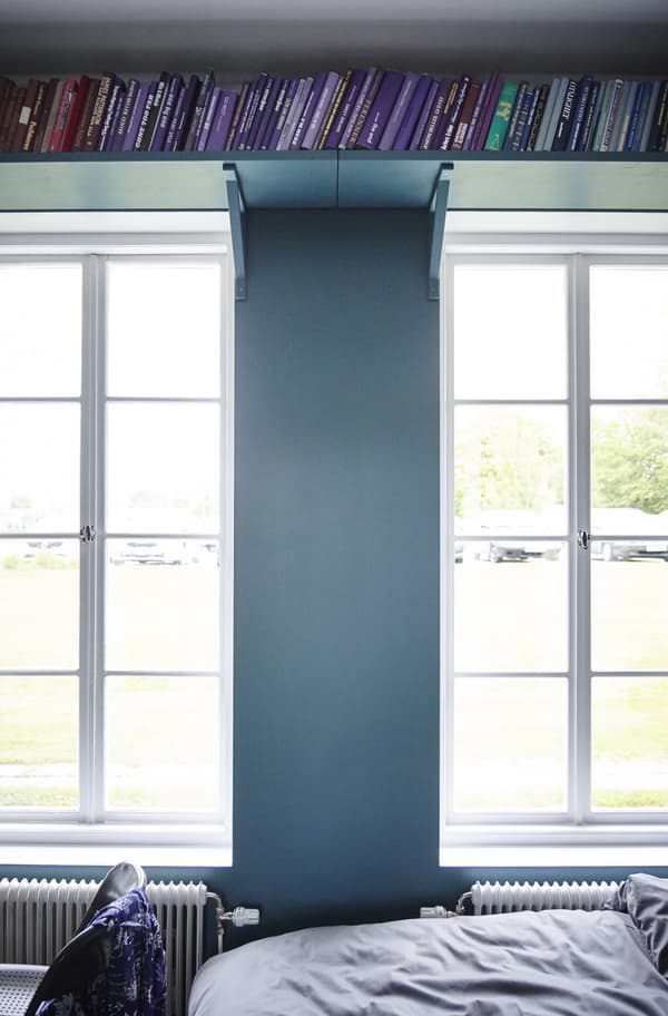 Wall-mounted shelves hold books in the space above the windows in a blue bedroom.