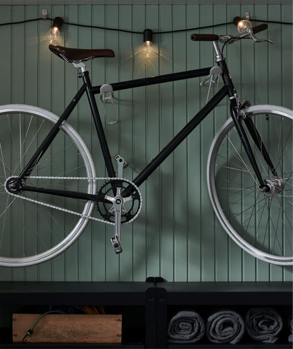 Wall section with a bike mounted on hooks and lit with a lighting chain running along the wall. A shelving unit sits below.