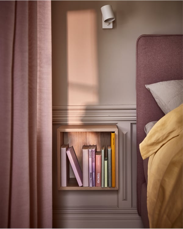 Wandgemonteerde open kast in wit gebeitst eikenpatroon, een witte wandlamp en een boxspring en gordijnen in roze.