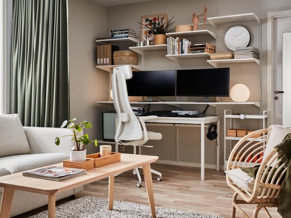 Wandregale, auf denen sich u. a. zwei Computerbildschirme und Bücher befinden; darunter stehen ein Couchtisch in Eschenfurnier und ein Drehstuhl in Beige/Weiß.