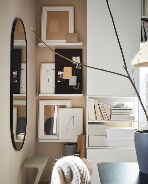 Wąska ściana z białymi półkami na zdjęcia. Leżą tu ciemnobrązowa tablica SVENSÅS i artystyczne ramki.