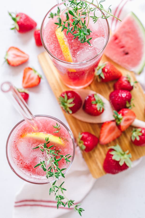 Watermelon-Strawberry Lemonade filled into two glasses on wooden tray decorated with strawberries and herbs 