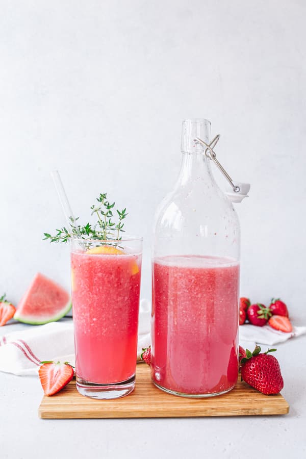 Watermelon-Strawberry Lemonade in glas next to a glas bottle with stopper on wooden tray 