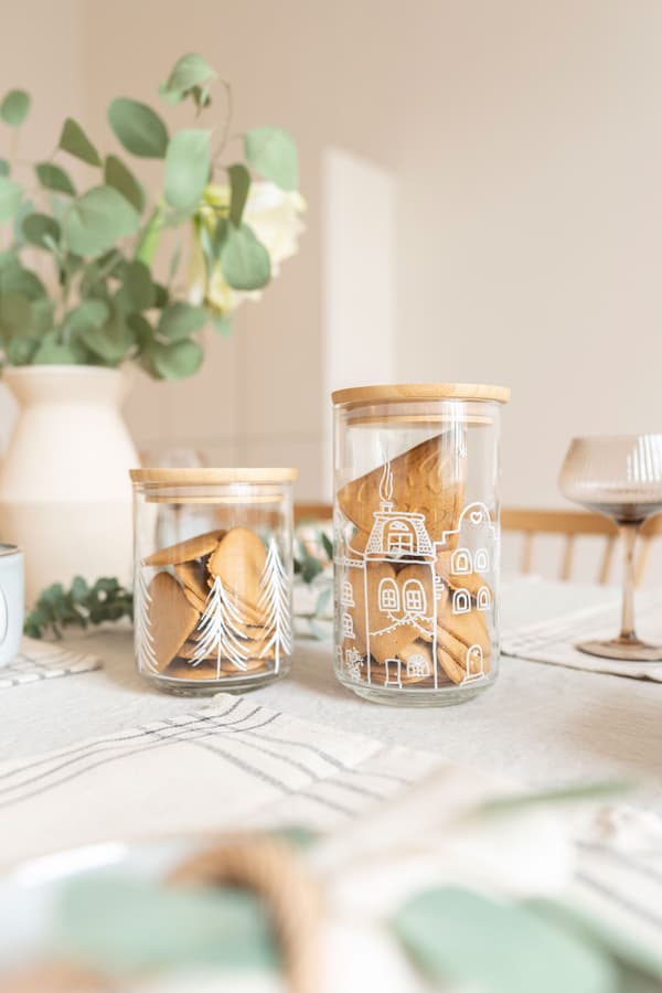 Weihnachtlich bemalte Gläser stehen auf einem Tisch mit weißer Tischdecke. Die selbstbemalten Vorratsgläser EKLATANT sind gefüllt mit Weihnachtsplätzchen.