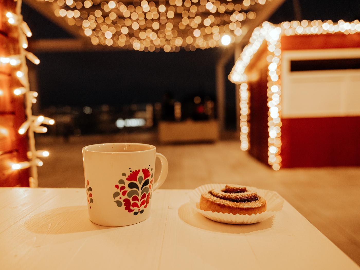 Weihnachtlich dekorierter Außenbereich mit Lichterketten, Zimtschnecke auf einem Teller und festlicher Tasse auf Holztisch – stimmungsvolle Atmosphäre auf einem Weihnachtsmarkt oder Winterevent.