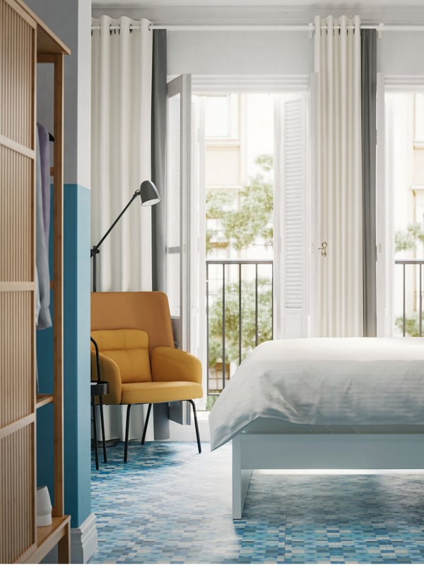 Weisses Bettgestell mit weisser Bettwäsche neben einem gelben Sessel in einem hellen Hotelzimmer.