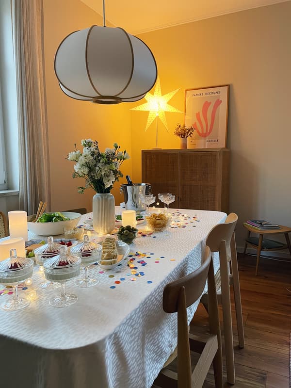 Weite Portraitaufnahme der geschmückten Silvestertafel mit weißer Tischdecke, buntem Konfetti und den kristallenen Sekt- und Dessertgläsern sowie der stimmungsvollen LED Beleuchtung.