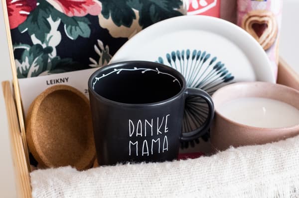 Weitere Nahaufnahme des Geschenkkorbes mit seinem Inhalt: eine Tasse, eine Duftkerze, ein Dessertteller, eine Kissenhülle.
