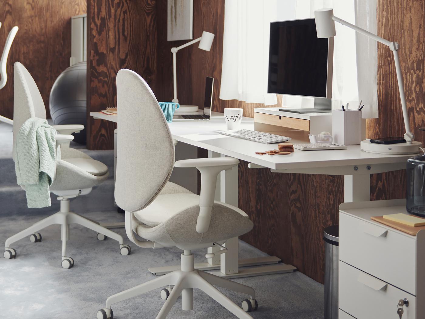 Werkplekken met witte TROTTEN bureaus en HATTEFJÄLL en LÅNGFJÄLL bureaustoelen in een bruine kantoorruimte met tapijt en houten wandbekleding.
