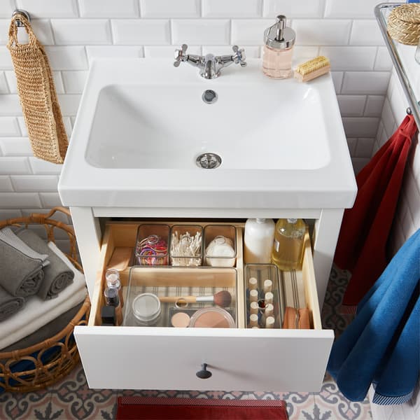وحدة حوض غسل بيضاء مع درج مفتوح وداخله صناديق صغيرة داكنة مع أغطية تُخزّن أغراض الحمام الصغيرة، مثل الماكياج.