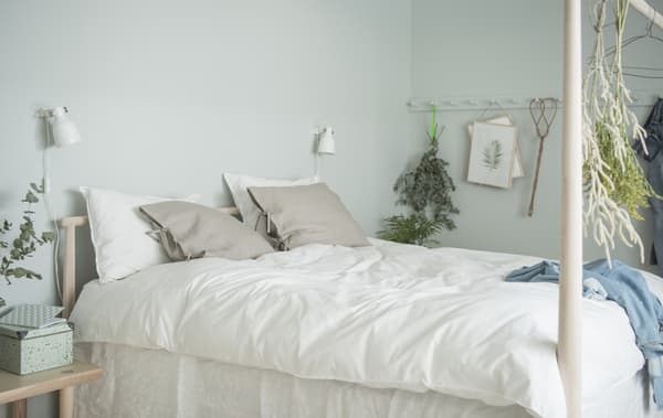 White and beige textiles in GJÖRA birch bed with a low headboard and high footboard shown together with two wall spotlights.