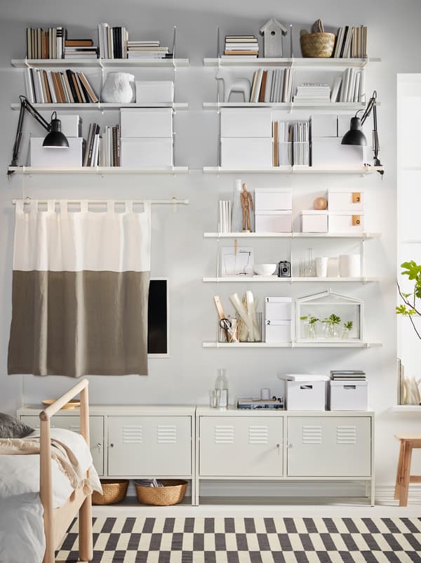 White BERGSHULT/PERSHULT shelves, filled with storage and decorations, and white IKEA PS cabinets cover a bedroom wall.