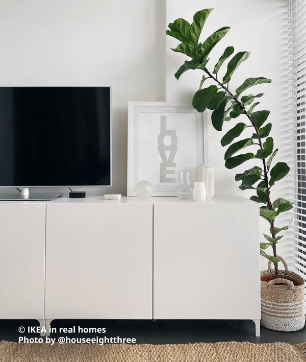 White BESTA TV console combination in the living room with push-to-open doors, next to a tall green plant with a natural fibre plant pot. A large television, a white frame with a poster and some vases and decorative items are also placed on top of the TV bench. Photo by instagrammer @houseeightthree for IKEA in real homes. 