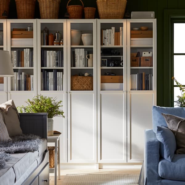 White BILLY bookcases with panel/glass doors, filled with books, storage boxes and curios, next to a window in a living room.