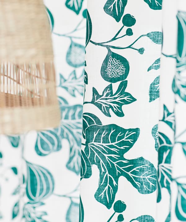 White curtain with a green pattern of fig tree branches, shown next to a ceiling lamp of natural banana fibre.