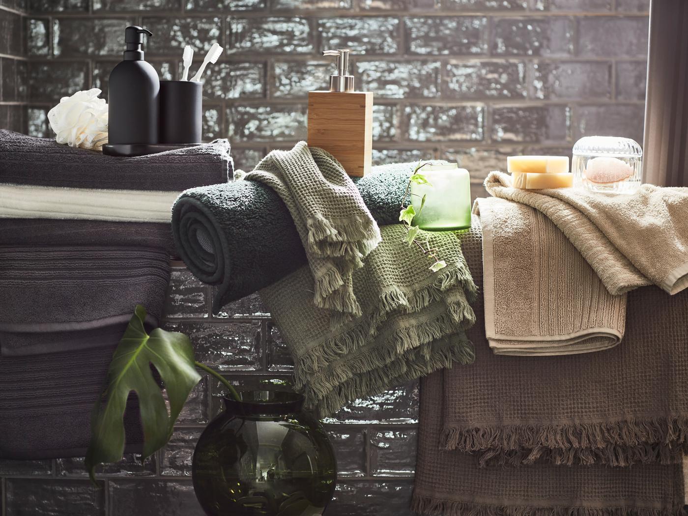 White, light beige, light grey/brown, dark grey and green towels displayed with various accessories in a grey-tiled bathroom.