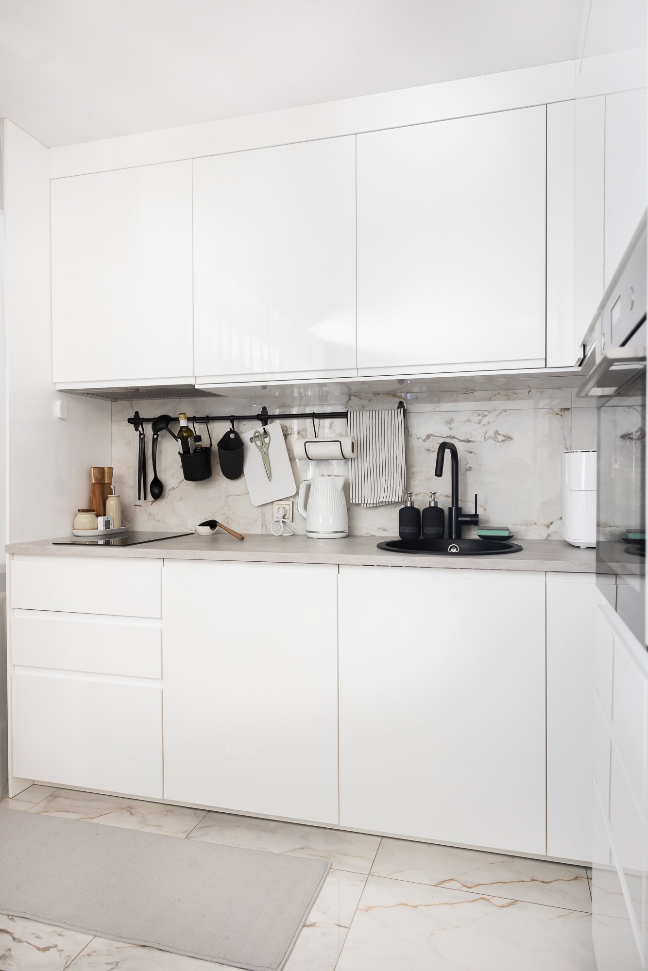 White modern kitchen with lower and upper cabinets reaching the ceiling, a built-in sink and a wall rail with hanging kitchen utensils.