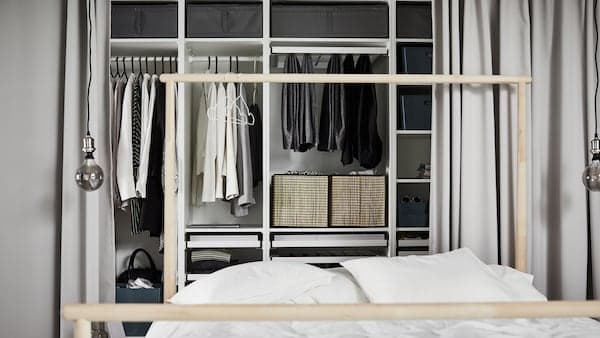 White PAX wardrobe combination containing different sections with multiple clothing items behind a bed with white duvet.