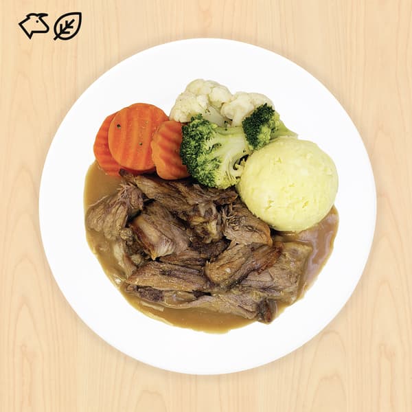 White plate of tender sliced roast beef in brown gravy, with mashed potatoes, carrots, broccoli, and cauliflower on a wood table.
