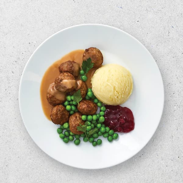 White plate on grey concrete background with IKEA meatballs, mash potato, peas, gravy, and jam