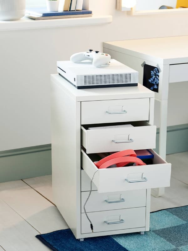 White room where a light HELMER drawer unit with colourful labels stands by a wooden post with a skateboard leaning on it.