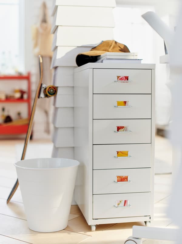 White room where a light HELMER drawer unit with colourful labels stands by a wooden post with a skateboard leaning on it.