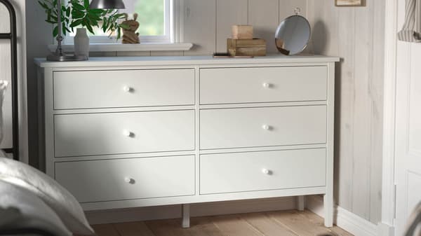 White six-drawer dresser with lamp, mirror and plant in a bright, cozy bedroom.