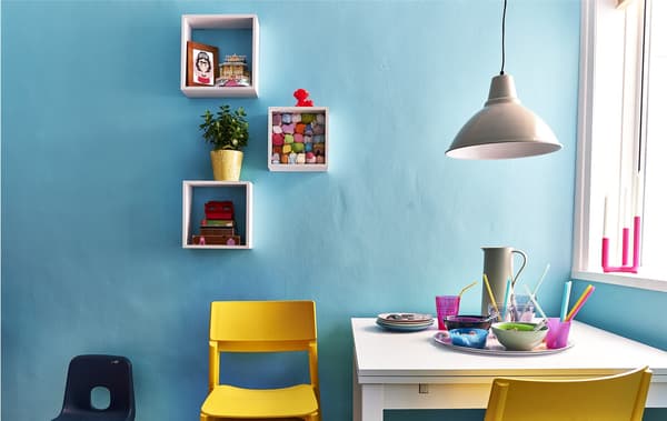 White square-shaped wall cubes with keepsakes above a dining area with white extendable table and a stack of yellow chairs.