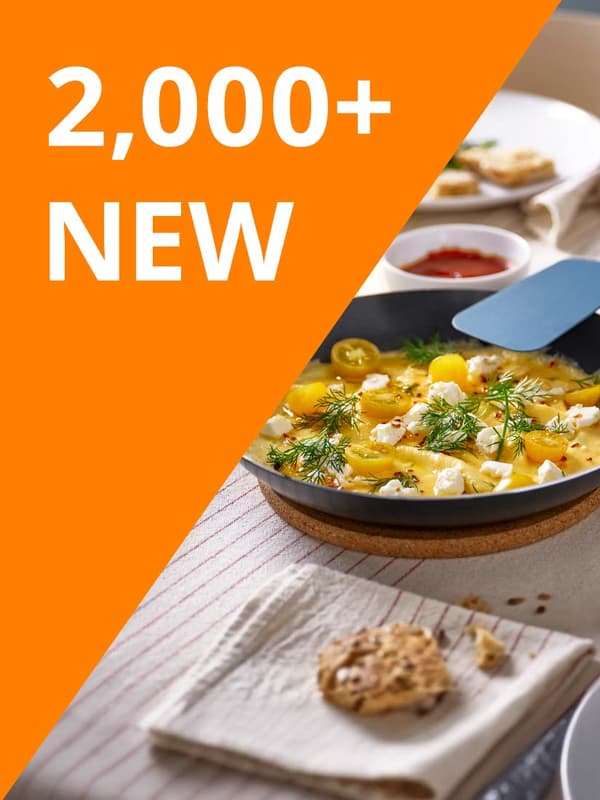 White text reads: "2,000+ NEW" on orange background. Black TAGGHAJ frying pan filled with food resting on a pot holder on dining table.