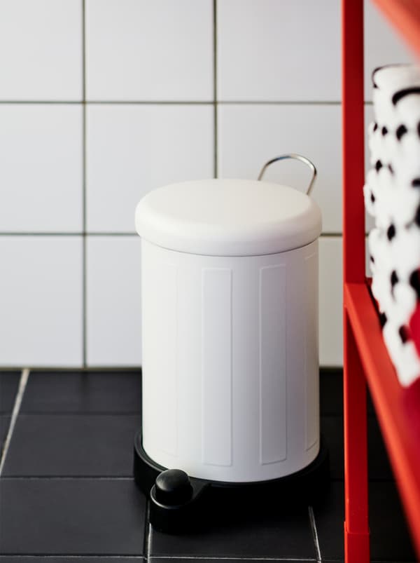White TOFTAN waste bin standing in a white-walled, black-floored, tiled bathroom next to a red open-storage ENHET shelving unit.