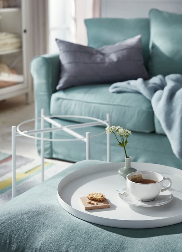 White tray taken off IKEA GLADOM white tray table and placed on a green sofa footstool, holding a teacup and a small sweet cracker.