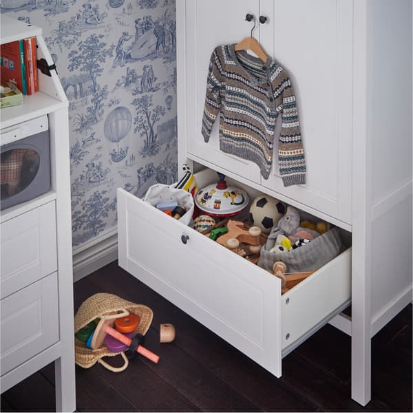 White wardrobe with a bottom drawer that's open and shows stored toys. A children's sweater hangs above on the door's knob.