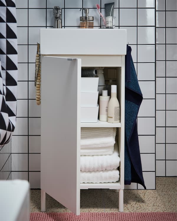 White wash basin cabinet with towels inside, a pink bath mat on the floor and a dark blue towel on the side of the cabinet.