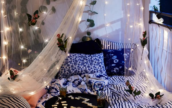 Wide bed on an enclosed balcony, its mosquito net decorated with flowers and LED lighting chains; drinks and game on a tray.