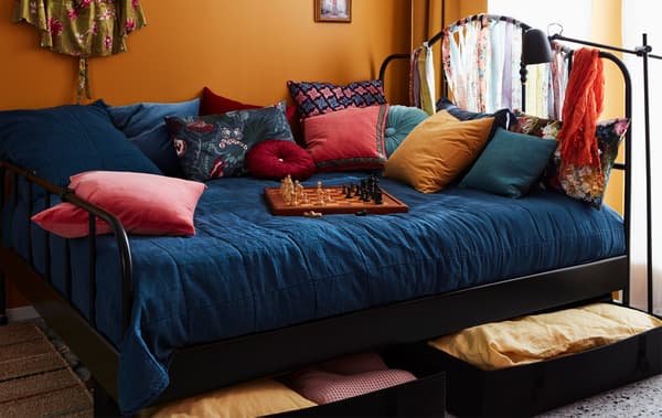 Wide, colourfully made bed with a lot of cushions and a game of chess on top. Bed linen stored in boxes underneath.