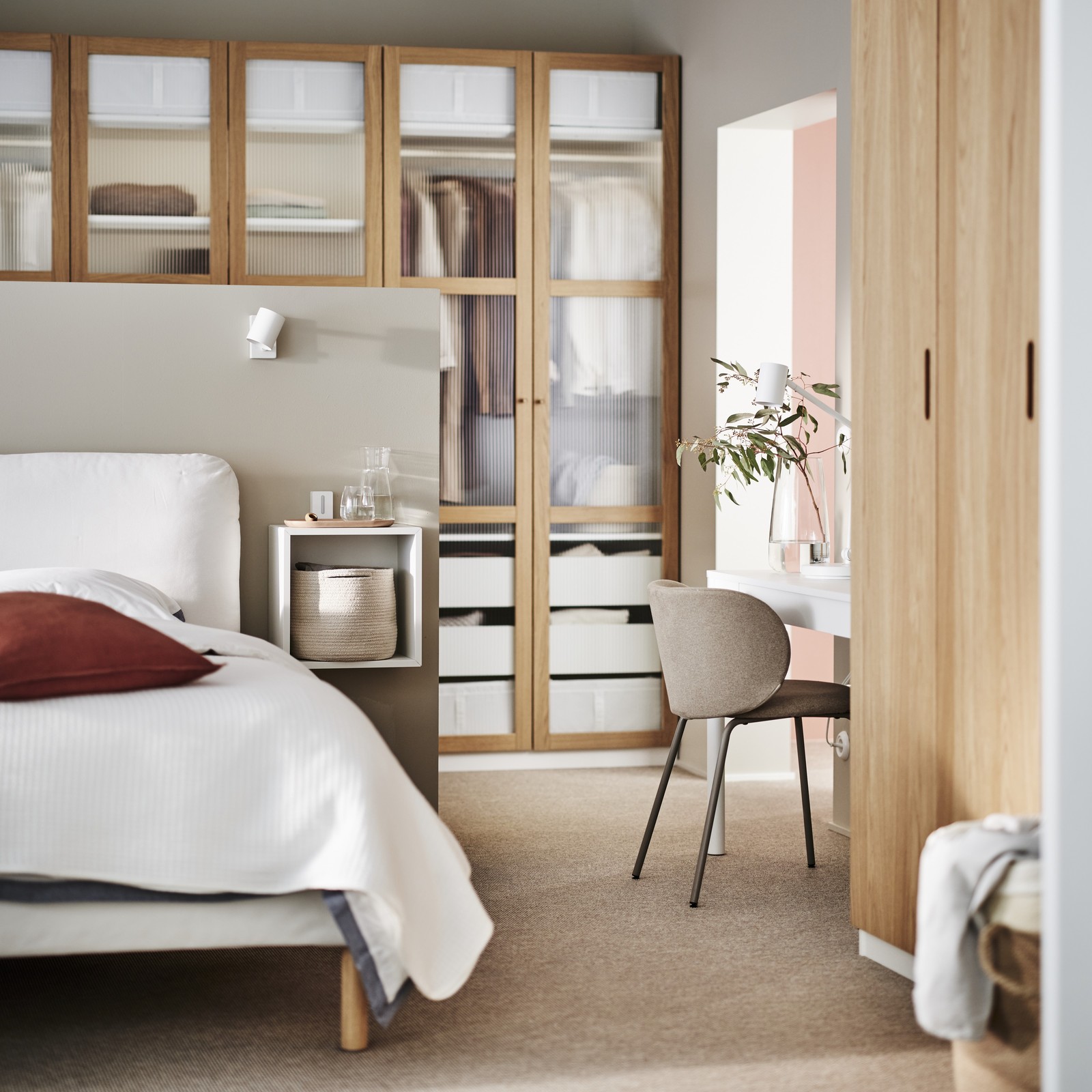 Widok pokoju na którym jest kawałeś łóżka z bialą pościelą i bordową poduszką, dębowe szafy, beżowe krzesło i beżowy dywan.