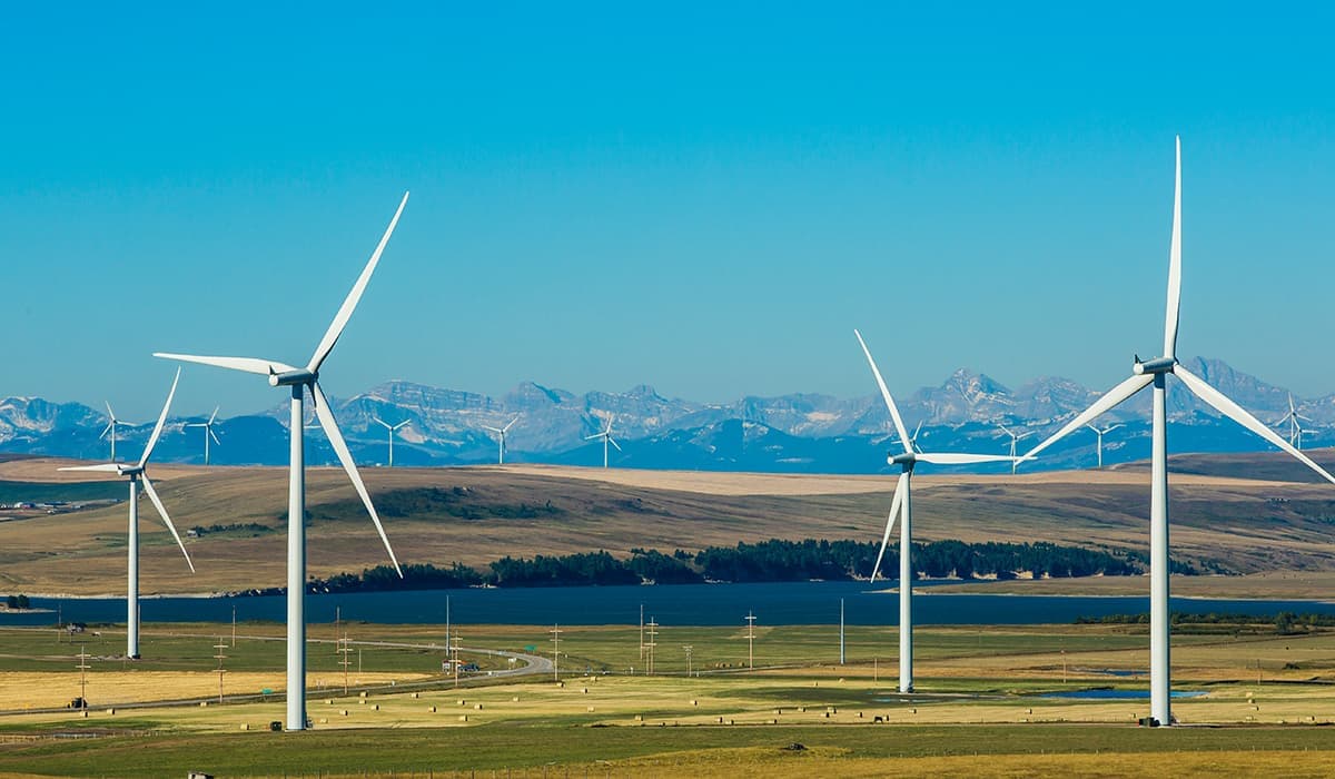 Windkraftanlagen ragen hoch in einer weiten, grasbewachsenen Landschaft empor, während ferne Berge und ein klarer blauer Himmel eine malerische Kulisse für erneuerbare Energien bilden.