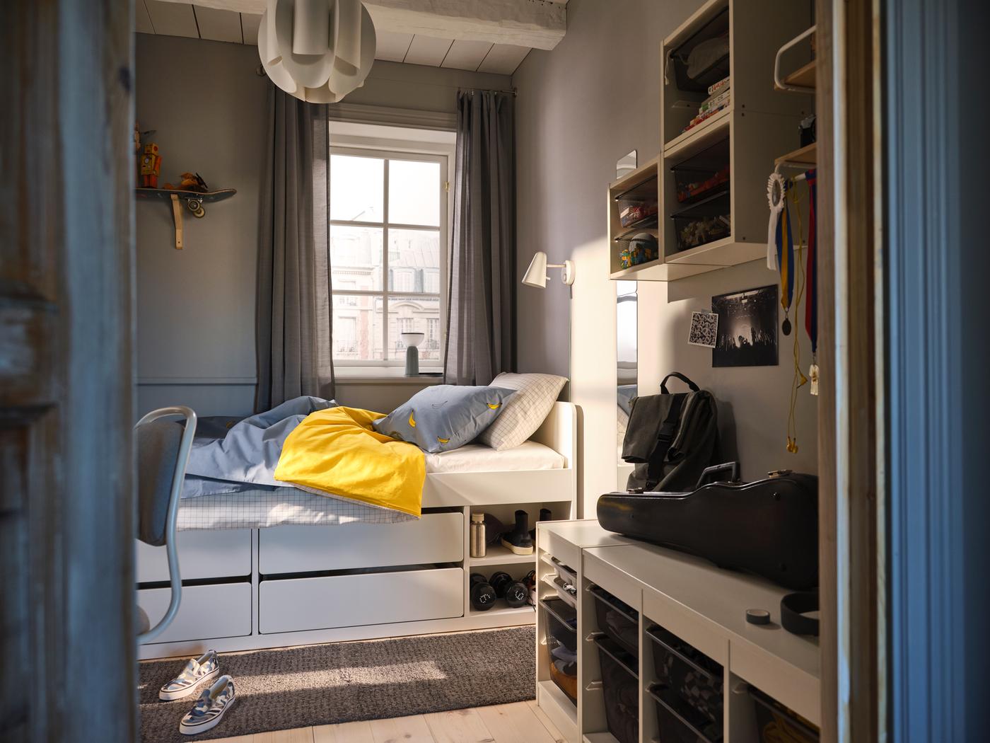 Wit SLÄKT bed met opbergruimte staat voor een raam in een kleine slaapkamer met TROFAST opbergers aan de wand en op de grond.