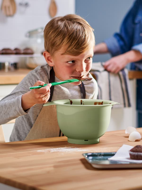 With a green/white spatula, a child in an apron eats chocolate cake batter from a green mixing bowl.