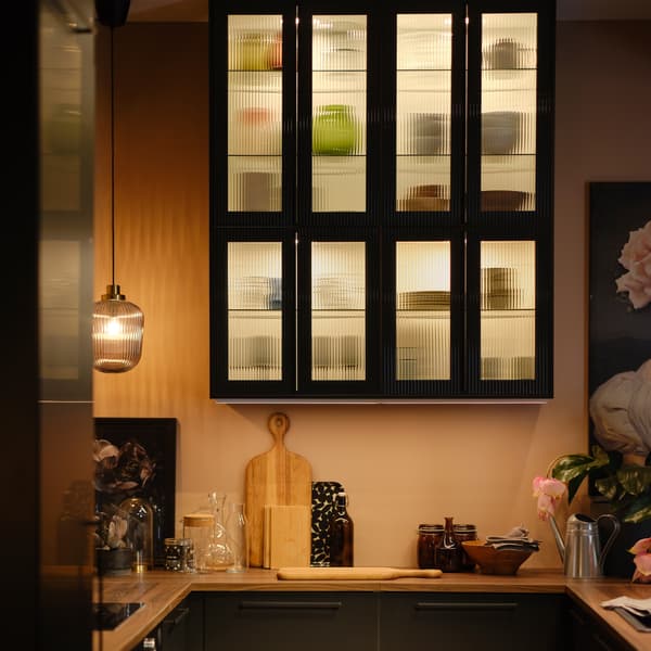 Witte METOD wandkasten met kastverlichting, legplanken en HEJSTA deuren in antraciet met getextureerd glas waarin diverse voorwerpen staan.