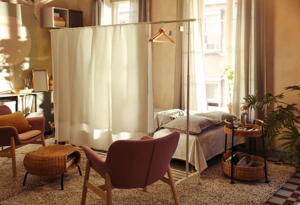 Wohnraumeinrichtung, bei der ein großes Bett hinter einem Raumteiler aus einem RIGGA Garderobenständer und VÅRELD Tagesdecken steht