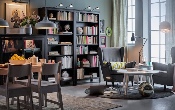 Wohnzimmer mit dunklem Bücherregal, einem Ohrensessel und Esstisch und zu jedem Bereich die passende Beleuchtung.